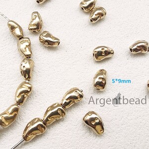 Pode incluir: Contas de metal douradas com uma superfície texturizada. As contas são enfiadas em um fio prateado. As contas medem 5 mm por 9 mm. O texto "Argentbead" é visível na imagem.