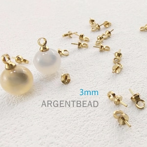 以下が含まれることがあります： 金色の金属製のビーズキャップで、上部にループが付いています。ビーズキャップは3mmのサイズで、白い表面に置かれています。画像には「3mm ARGENTBEAD」というテキストも表示されています。