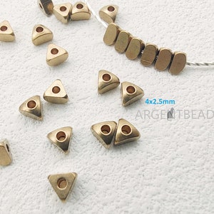Puede incluir: Cuentas triangulares doradas con un agujero en el centro. Las cuentas miden 4 mm x 2,5 mm. Las cuentas están esparcidas sobre una superficie blanca. El texto "4x2,5 mm ARGENITEBEAD" es visible en la imagen.