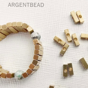May include: A gold bracelet made of square-shaped beads with two green and white stones. The bracelet is surrounded by small gold metal pieces with holes in them.