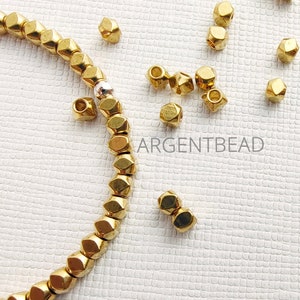 Pode incluir: Close-up de contas douradas e facetadas espalhadas sobre uma superfície branca texturizada. Algumas contas são enfiadas, formando um colar ou fio. O texto "ARGENTBEAD" é visível na imagem.