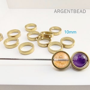 May include: A group of 10mm gold-toned metal rings with two holes each. The rings are scattered on a white surface. There are two round beads, one with a purple stone and one with a clear stone with orange flecks, on a silver rod.