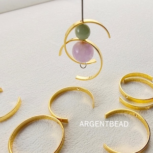 以下が含まれることがあります： 2つの穴が空いた金色の金属製の輪が5つと、ピンクと緑の石が取り付けられた金色の金属製の輪が1つ。画像には「ARGENTBEAD」というテキストが表示されています。