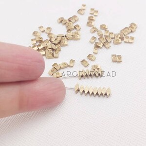 May include: A collection of small, gold-colored metal beads in various shapes, including small squares and elongated ovals. Some beads are strung on a thin wire. The image includes the text "ARGENTBEAD".