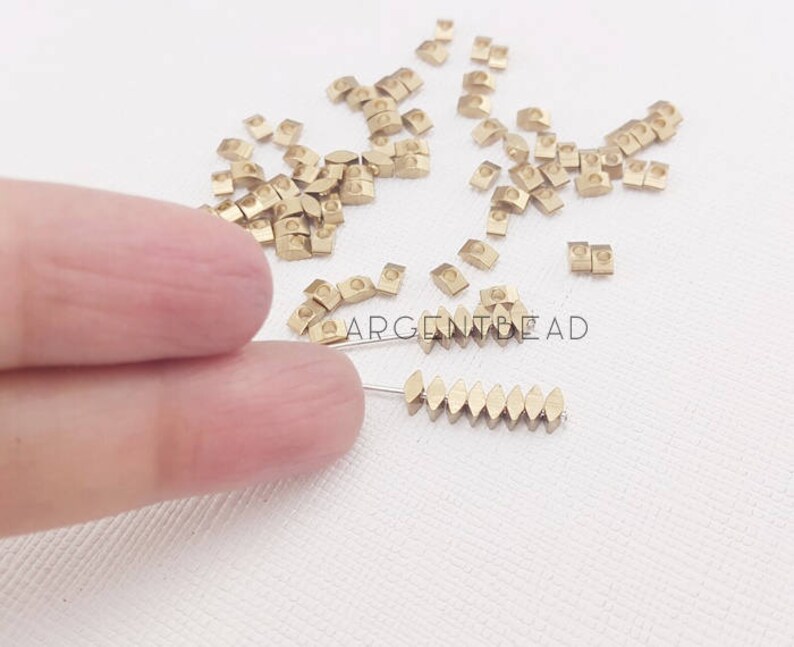 May include: A collection of small, gold-colored metal beads in various shapes, including small squares and elongated ovals. Some beads are strung on a thin wire. The image includes the text "ARGENTBEAD".