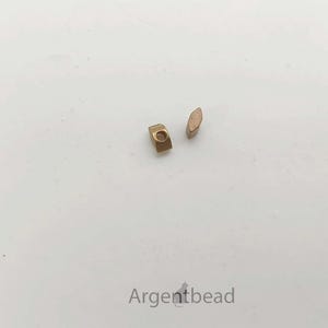 May include: Two small, gold-colored metal beads on a white surface. One bead is a small rectangle with a hole, and the other is an oval shape. The beads are likely for jewelry making or crafting.