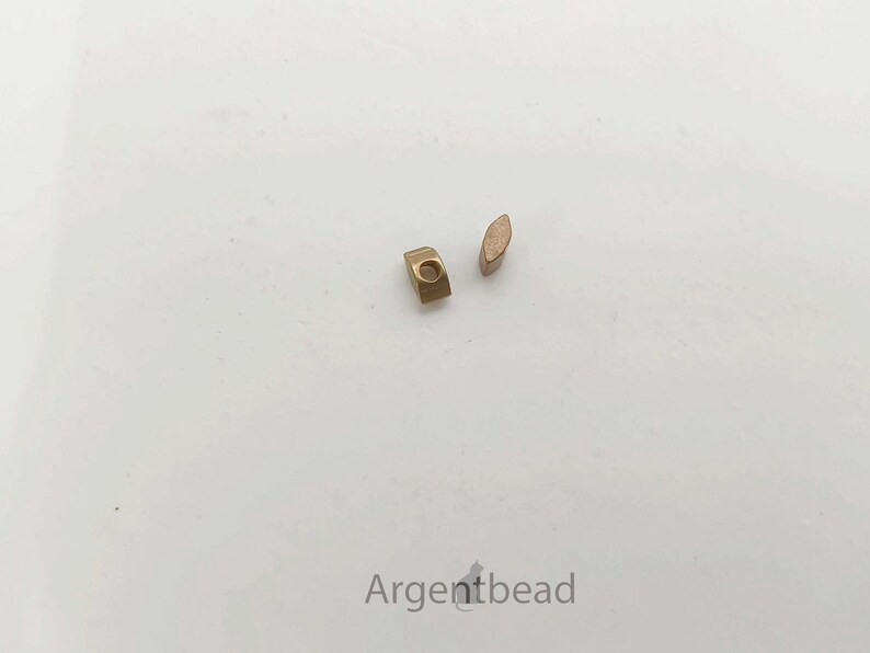 May include: Two small, gold-colored metal beads on a white surface. One bead is a small rectangle with a hole, and the other is an oval shape. The beads are likely for jewelry making or crafting.