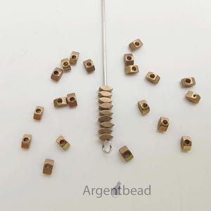 May include: A collection of small, bronze-colored metal beads scattered on a white surface. Some beads are square with a hole in the center, while others are oval-shaped and strung together. The text "Argentbead" is visible.