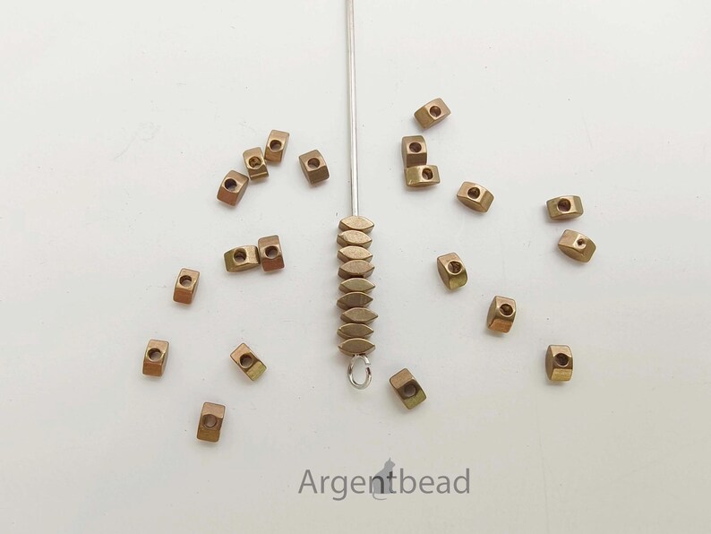 May include: A collection of small, bronze-colored metal beads scattered on a white surface. Some beads are square with a hole in the center, while others are oval-shaped and strung together. The text "Argentbead" is visible.