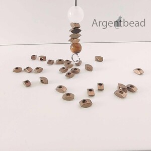 May include: A collection of small, metallic-colored beads scattered on a white surface. A strand of beads hangs from above, featuring a white sphere, stacked geometric shapes, and a brown bead. The word "Argentbead" is visible in the upper right corner.