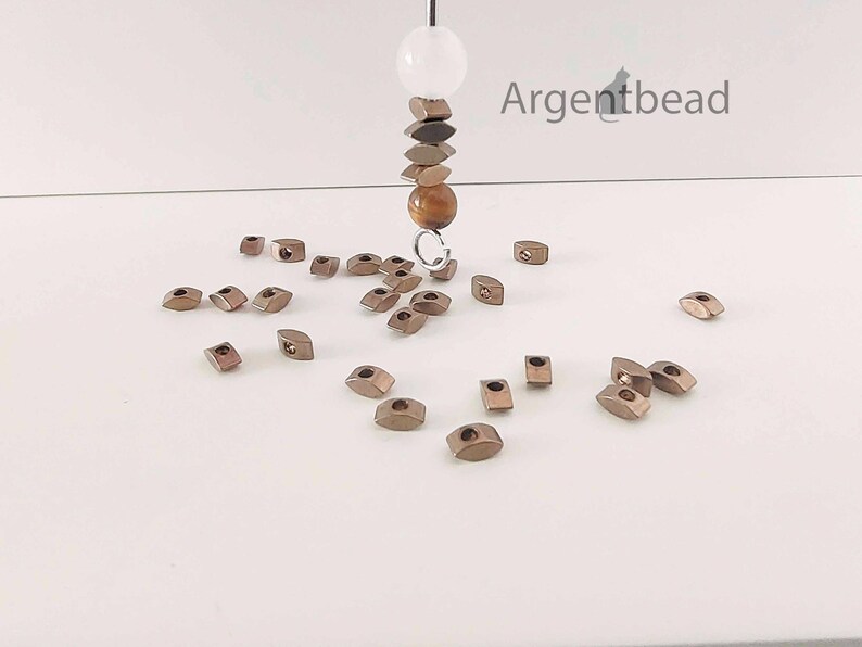 May include: A collection of small, metallic-colored beads scattered on a white surface. A strand of beads hangs from above, featuring a white sphere, stacked geometric shapes, and a brown bead. The word "Argentbead" is visible in the upper right corner.