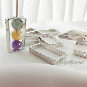 May include: A silver rectangular pendant with three colorful gemstone beads: green, yellow, and purple. Several empty silver rectangular frames are scattered around the pendant, suggesting a jewelry-making theme.
