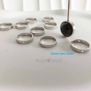 May include: A collection of silver-colored rings with a flat design and two small holes. The rings are approximately 13mm in diameter. A pendant with a dark stone is also visible. The text "13mm two holes" and "Argentbead" are present.