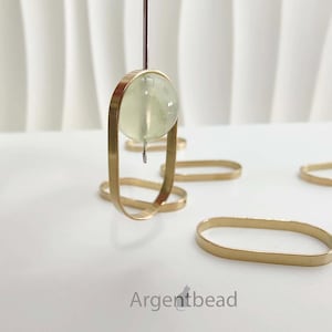 May include: A pale green translucent spherical bead suspended on a thin metal needle within a gold-toned oval metal frame. Several additional empty gold-toned oval metal frames are scattered nearby. The text Argentbead is printed at the bottom.