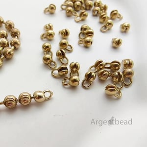 May include: Close-up of gold-colored bead caps with small loops, scattered on a white surface. Some are strung together, while others are loose. The bead caps have a textured, rounded design, perfect for jewelry making.