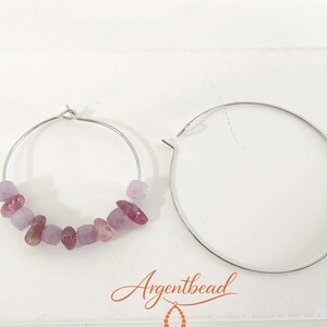 Può includere: Un paio di orecchini a cerchio color argento. Un cerchio è semplice, mentre l'altro è decorato con una serie di piccole perline di pietre preziose irregolari, di colore viola chiaro e rosa scuro. Il testo Argentbead è stampato in arancione.