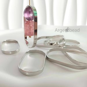 Op de afbeelding: Een collectie zilverkleurige metalen ovale frames voor het maken van sieraden, met een gladde, gepolijste afwerking. Eén frame hangt verticaal en bevat een doorschijnende paarse bolvormige kraal en een getextureerde roze cilindrische kraal.
