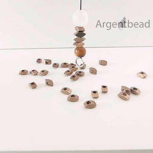 May include: An assortment of small, metallic-colored beads scattered on a white surface. A strand of beads hangs from above, featuring a white sphere, brown and bronze-toned beads, and a round, amber-colored bead. The Argentbead logo is visible in the upper right corner.