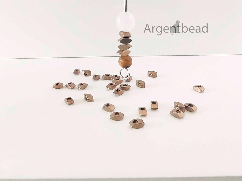 May include: An assortment of small, metallic-colored beads scattered on a white surface. A strand of beads hangs from above, featuring a white sphere, brown and bronze-toned beads, and a round, amber-colored bead. The Argentbead logo is visible in the upper right corner.