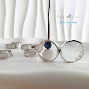 Może przedstawiać: Srebrne pierścienie z jednym otworem, niektóre z niebieską i białą perłą w środku. Pierścienie są prezentowane na białej powierzchni. Widoczny jest tekst "Argentbead 15mm One hole".
