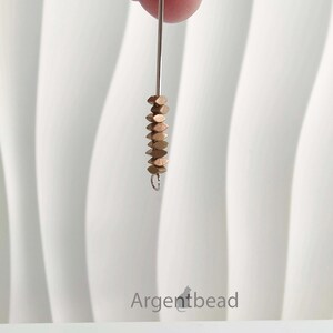 May include: A strand of small, copper-colored, flat beads suspended from a silver-colored wire. The beads are stacked closely together, creating a textured effect. The background is a white, textured surface.