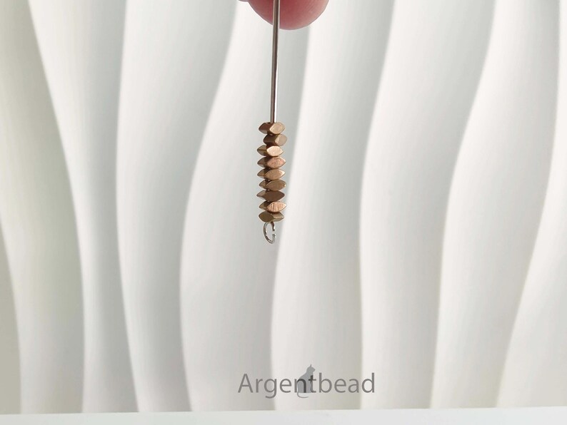 May include: A strand of small, copper-colored, flat beads suspended from a silver-colored wire. The beads are stacked closely together, creating a textured effect. The background is a white, textured surface.