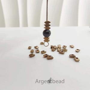 May include: A close-up of jewelry-making supplies. A dark bead is suspended between stacked, bronze-colored, diamond-shaped beads. Scattered around are more bronze-colored diamond-shaped beads. The word "Argentbead" is in the bottom right corner.