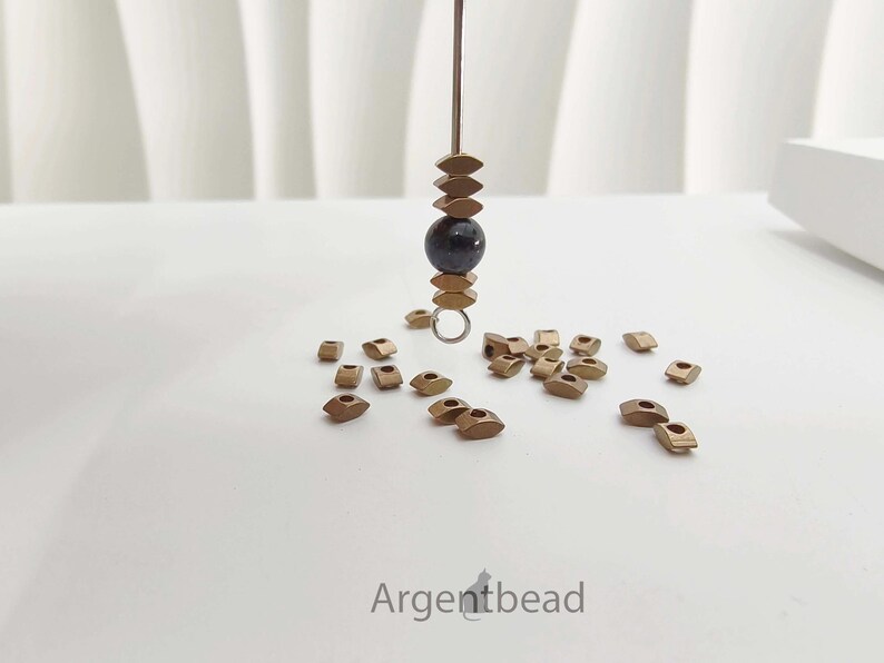 May include: A close-up of jewelry-making supplies. A dark bead is suspended between stacked, bronze-colored, diamond-shaped beads. Scattered around are more bronze-colored diamond-shaped beads. The word "Argentbead" is in the bottom right corner.
