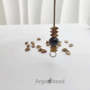 May include: A close-up of a jewelry component with a dark blue bead, gold-colored metal beads, and a small silver ring. The component is suspended on a metal rod. Scattered around are more gold-colored metal beads. The word "Argentbead" is at the bottom.
