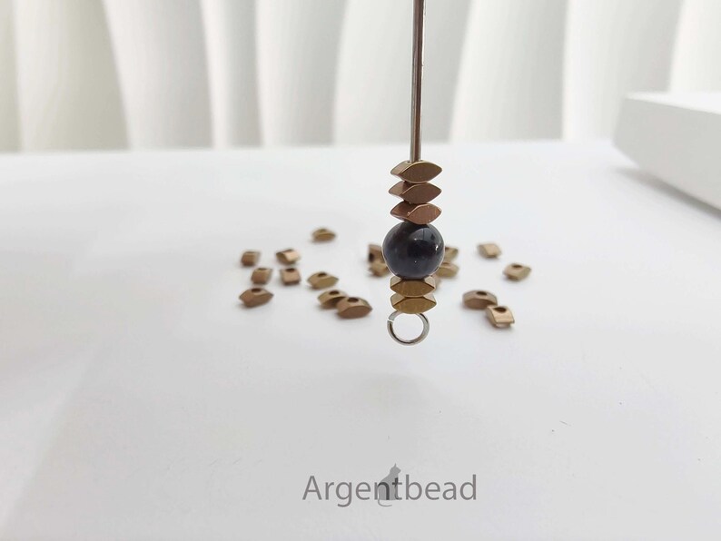 May include: A close-up of a jewelry component with a dark blue bead, gold-colored metal beads, and a small silver ring. The component is suspended on a metal rod. Scattered around are more gold-colored metal beads. The word "Argentbead" is at the bottom.