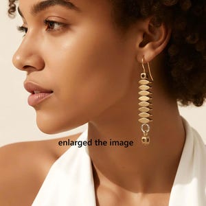 May include: Gold-toned dangle earrings. Each earring features a series of flat, oval-shaped elements stacked vertically, connected by small rings. A small, cube-shaped charm hangs at the bottom. The earrings are worn by a person.