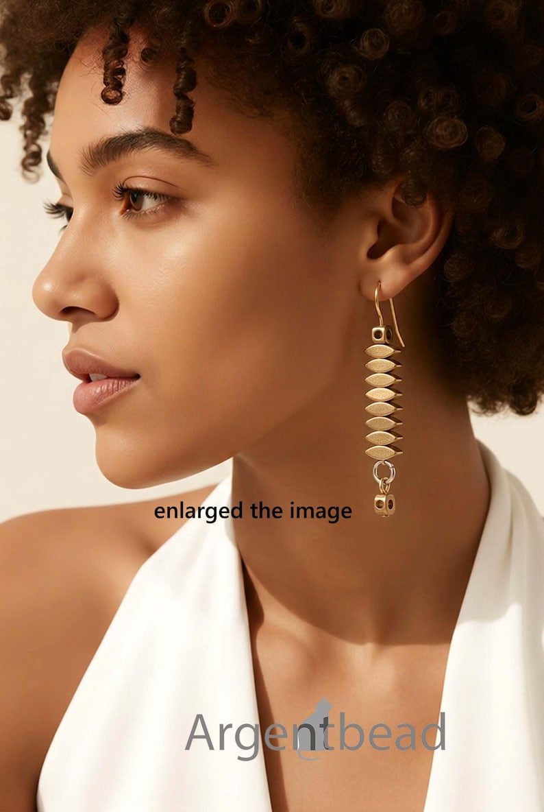May include: Gold-toned dangle earrings. Each earring features a series of flat, oval-shaped elements stacked vertically, connected by small rings. A small, cube-shaped charm hangs at the bottom. The earrings are worn by a person.