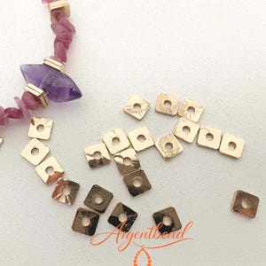 Könnte beinhalten: Eine Sammlung kleiner, quadratischer, goldfarbener Metallperlen mit einem Mittelloch, verstreut neben einem Teilstrang aus rosa Splitterperlen und einer einzelnen violetten, marquiseförmigen Steinperle mit goldfarbenen Metallzwischenstücken.