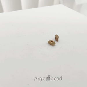 May include: Two small, gold-colored metal beads are displayed on a white surface. One bead is a rounded rectangle with a hole, and the other is a small, angled rectangle. The word "Argentbead" is at the bottom.