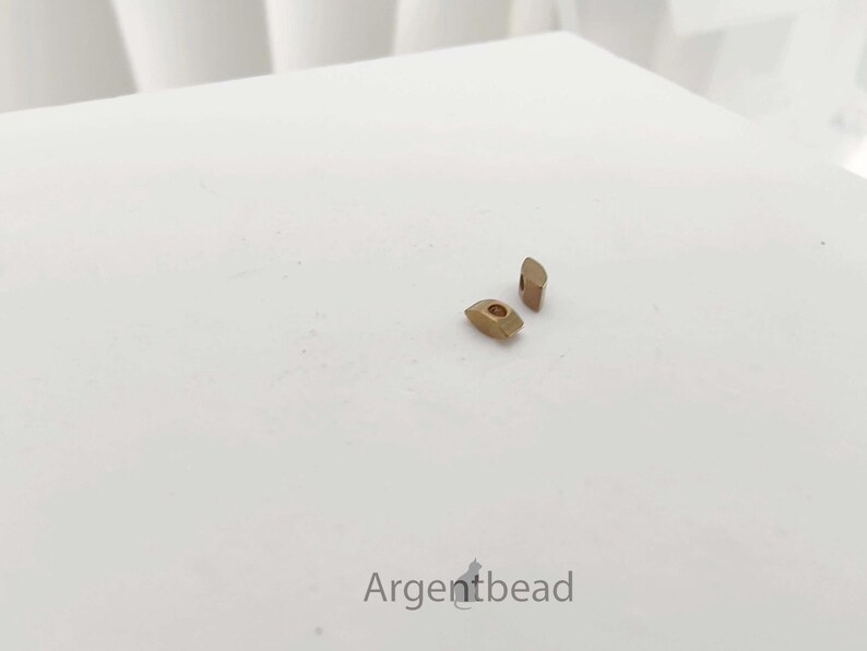 May include: Two small, gold-colored metal beads are displayed on a white surface. One bead is a rounded rectangle with a hole, and the other is a small, angled rectangle. The word "Argentbead" is at the bottom.