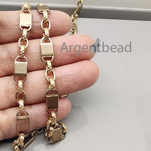 Può includere: Collana a catena color oro con maglie rettangolari e ovali. La catena è tenuta in mano, con il nome del marchio "Argentbead" visibile. La collana presenta un design strutturato.