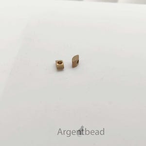 May include: Two small, gold-colored metal beads with a square and half-circle shape are displayed on a white surface. The beads are positioned near each other, showcasing their design and texture. The image is well-lit, highlighting the details of the jewelry components.