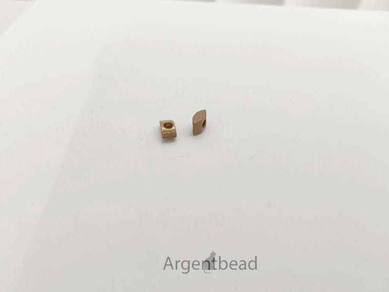 May include: Two small, gold-colored metal beads with a square and half-circle shape are displayed on a white surface. The beads are positioned near each other, showcasing their design and texture. The image is well-lit, highlighting the details of the jewelry components.