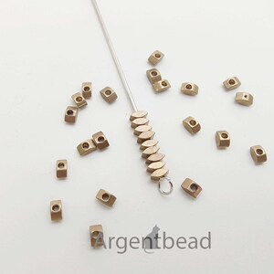 May include: A collection of small, metallic-colored beads scattered on a white surface. The beads are rectangular with a hole through the center. A silver needle with a loop and a string of similar beads is also present. The Argentbead logo is visible.