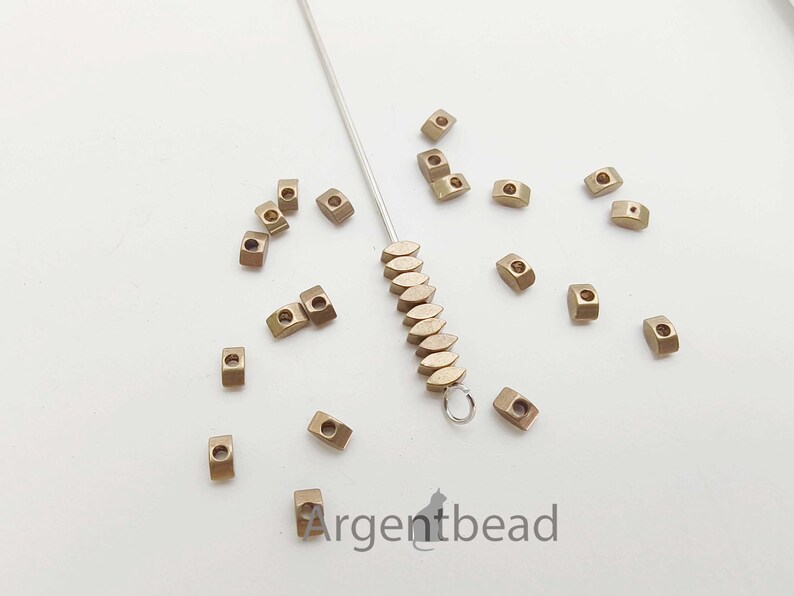 May include: A collection of small, metallic-colored beads scattered on a white surface. The beads are rectangular with a hole through the center. A silver needle with a loop and a string of similar beads is also present. The Argentbead logo is visible.