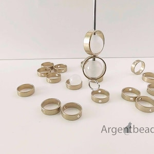 May include: Gold-colored metal rings with various designs, including some with small cut-out shapes. Some rings are arranged around a central display featuring two large clear orbs. The rings are scattered on a white surface.