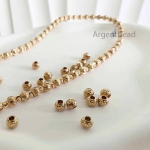 May include: Gold-colored metal beads, some strung together, scattered on a white surface. The beads have a detailed floral design and are likely for jewelry making. The brand name "Argenthead" is visible in the upper right corner.