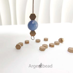 Puede incluir: Primer plano de componentes de joyería, que incluyen pequeñas cuentas hexagonales de color dorado dispersas sobre una superficie blanca. Un hilo de cuentas presenta una esfera azul y dos cuentas hexagonales. El logotipo de Argentbead es visible.