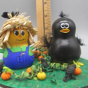 May include: Hand-painted gourd figurines of a scarecrow and a black bird, with a pumpkin and crow theme. The scarecrow wears a straw hat and blue overalls. The bird has a yellow beak. The base is green with miniature pumpkins and crows.