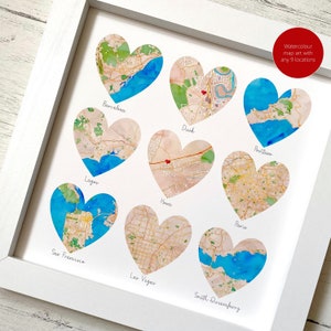 May include: A framed watercolour map art print displaying nine heart-shaped maps of locations like Barcelona and Dusk. The maps use beige, green, and blue hues, with location names beneath each heart. The white frame surrounds the artwork. The text reads: Watercolour map art with any 9 locations.