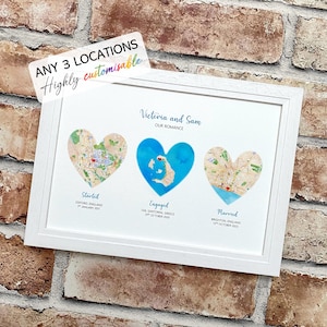 May include: A white framed print with three heart-shaped map illustrations. The print is personalised with the names "Victoria and Sam" and the text "Our Romance." The locations are labelled "Started," "Engaged," and "Married," with dates and places.