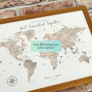 以下が含まれることがあります： 木製のフレームに入った世界地図。薄茶色と白を基調とし、黒いピンで場所を示しています。「LIFE IS A JOURNEY best travelled together」の文字が見えます。「Over 20 background colour options」の文字も表示されています。