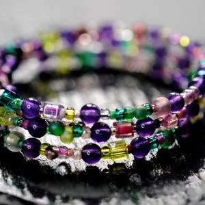 Op de afbeelding: Een meerstrengige armband gemaakt van paarse, groene en heldere kralen. De armband is om zichzelf gewikkeld en heeft een verscheidenheid aan verschillende kralen.