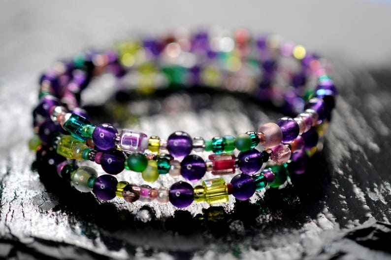 Op de afbeelding: Een meerstrengige armband gemaakt van paarse, groene en heldere kralen. De armband is om zichzelf gewikkeld en heeft een verscheidenheid aan verschillende kralen.