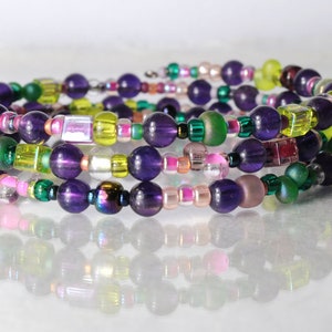Op de afbeelding: Een meerstrengige armband met paarse, groene, roze en gele kralen. De armband is gemaakt van verschillende maten en vormen kralen, waaronder ronde, vierkante en rechthoekige.